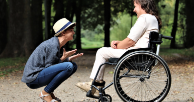 We Need To Talk More About Sex And Disability person wearing a white hat and blue jeans is crouching in front of a person wearing all white in a wheelchair. Both people are laughing together in a forest